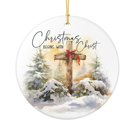 Round ceramic Christmas ornament featuring a watercolor winter scene with a wooden cross, snow-covered evergreen trees, and the text 'Christmas Begins With Christ' in black script, hanging from a gold string on a white background.