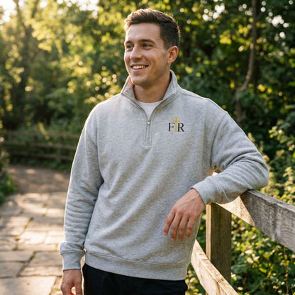 A man wearing a light gray heathered quarter-zip sweatshirt with a small black and gold embroidered logo on the left chest, standing outdoors on a wooden fence with a blurred green garden background.