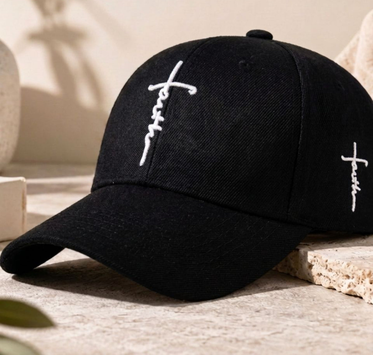 A black baseball cap with white embroidered cross designs on the front and side, resting on a textured beige stone surface with soft shadows.
